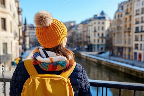 A woman faces away from the camera, showing an orange hat and multicolored scarf while standing on a bridge, set against an urban backdrop under a clear blue sky. photo