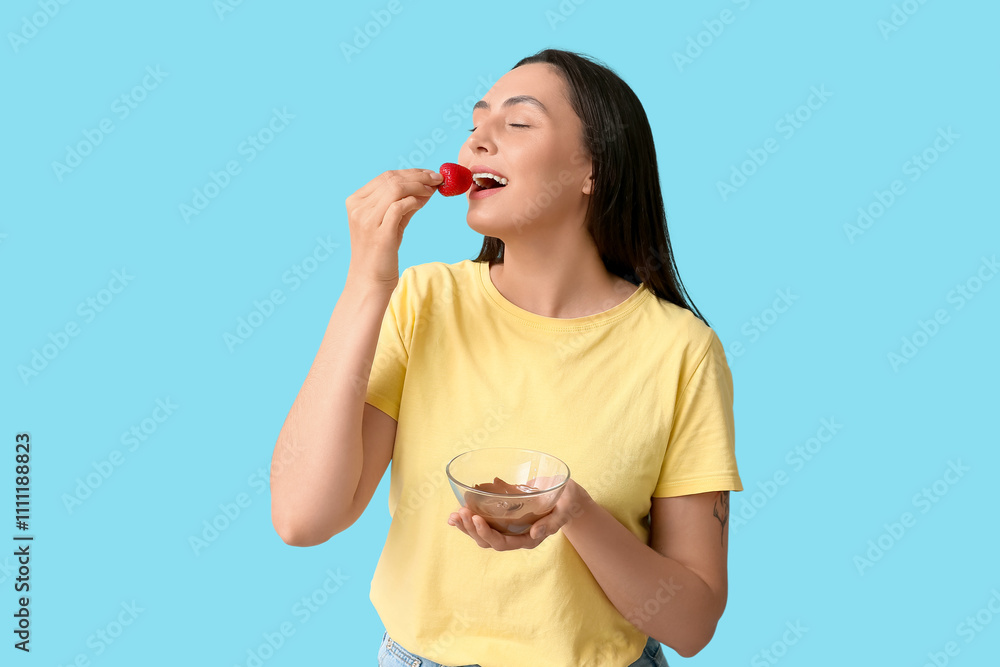 Beautiful young woman dipping fresh strawberry into bowl with melted chocolate on blue background Beautiful young woman dipping fresh strawberry into bowl with melted chocolate on blue background