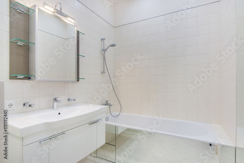 Laconic design of the bathroom decorated with white tiles. A cabinet with a mirror over the sink and a bathtub with a shower.