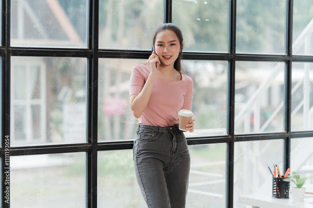 Smiling Businesswoman on Phone Call: A woman in a pink shirt and jeans stands by a large window, holding a cup of coffee and talking on her phone. She looks happy and confident. Smiling Businesswoman on Phone Call: A woman in a pink shirt and jeans stands by a large window, holding a cup of coffee and talking on her phone. She looks happy and confident.