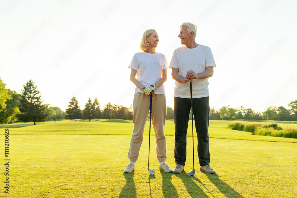happy elderly senior couple in white uniform holding golf clubs and hugging on golf course at sunset, old man and woman playing golf and smiling looking at camera in nature happy elderly senior couple in white uniform holding golf clubs and hugging on golf course at sunset, old man and woman playing golf and smiling looking at camera in nature
