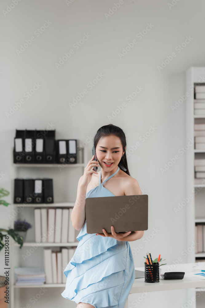 Businesswoman on the Go: A modern professional woman in a stylish blue dress seamlessly multitasks, holding a laptop and taking a call. Businesswoman on the Go: A modern professional woman in a stylish blue dress seamlessly multitasks, holding a laptop and taking a call.