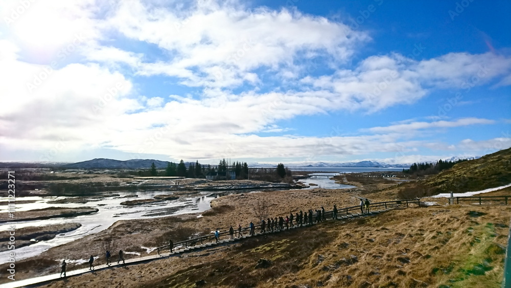 Iceland - 3.24.2018: Tourists walking down a staircase and a footpath towards frozen rivers among dried meadows in Thingvellir National Park under a cloudy blue sky with sunlight before the pandemic Iceland - 3.24.2018: Tourists walking down a staircase and a footpath towards frozen rivers among dried meadows in Thingvellir National Park under a cloudy blue sky with sunlight before the pandemic