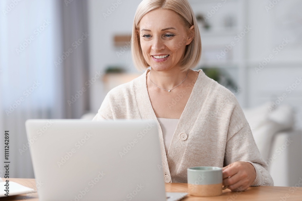 Happy middle aged woman with cup of drink using laptop at table indoors Happy middle aged woman with cup of drink using laptop at table indoors