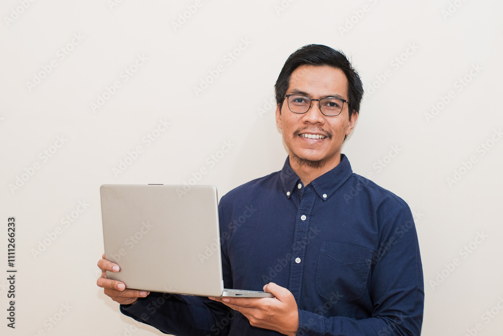 Smiling Southeast Asian man wearing blue shirt and glasses holding laptop, standing over isolated white background, Positive person Smiling Southeast Asian man wearing blue shirt and glasses holding laptop, standing over isolated white background, Positive person