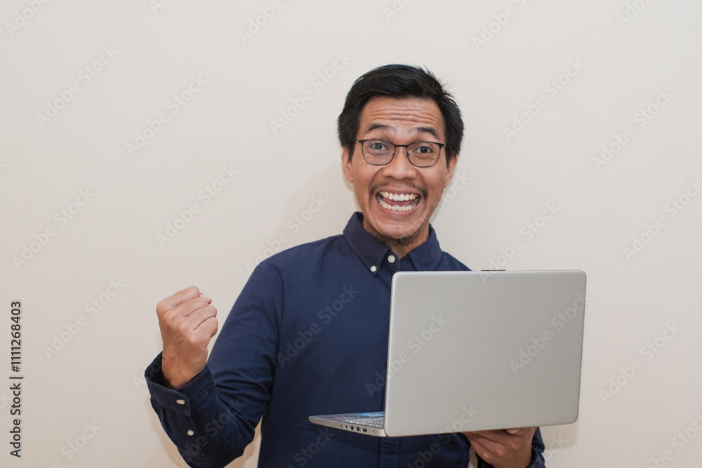 Smiling and satisfied Southeast Asian man wearing blue shirt with glasses holding laptop with hand shows winning victory gesture, standing over isolated white background, Positive person Smiling and satisfied Southeast Asian man wearing blue shirt with glasses holding laptop with hand shows winning victory gesture, standing over isolated white background, Positive person