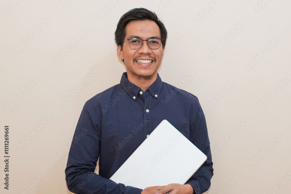 Smiling Southeast Asian man wearing blue shirt and glasses holding laptop, standing over isolated white background, Positive person Smiling Southeast Asian man wearing blue shirt and glasses holding laptop, standing over isolated white background, Positive person