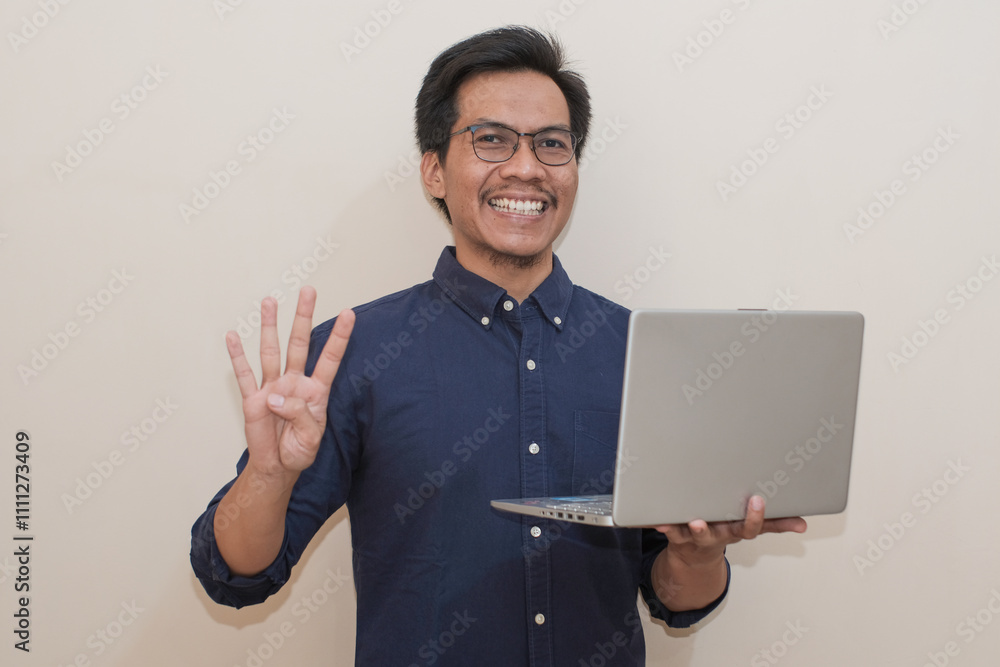 Smiling Southeast Asian man wearing blue shirt and glasses holding laptop with giving number 12345 by hand gesture , standing over isolated white background, Positive person Smiling Southeast Asian man wearing blue shirt and glasses holding laptop with giving number 12345 by hand gesture , standing over isolated white background, Positive person