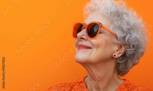 Happy smiling senior woman wearing sunglasses, orange solid colorful background, image with space for text / Femme âgée souriante et heureuse avec lunettes de soleil, fond orange, espace pour le texte