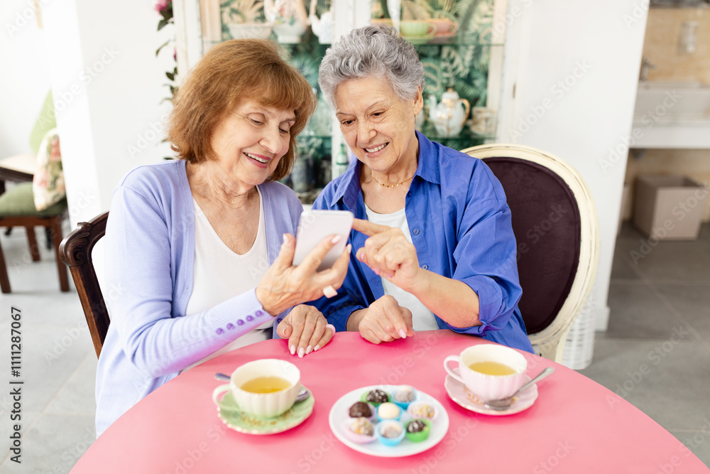 Two elderly women sit together at a round table enjoying dessert and tea. They are sharing laughter while looking at a mobile device. The cafe setting has a cozy ambiance. Two elderly women sit together at a round table enjoying dessert and tea. They are sharing laughter while looking at a mobile device. The cafe setting has a cozy ambiance.