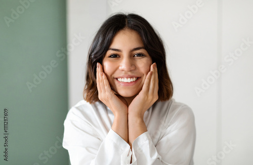 Closeup portrait of cheerful pretty young middle eastern woman in white silk robe touching her face and smiling, enjoying smooth glow moisturised skin, copy space. Face care routine concept