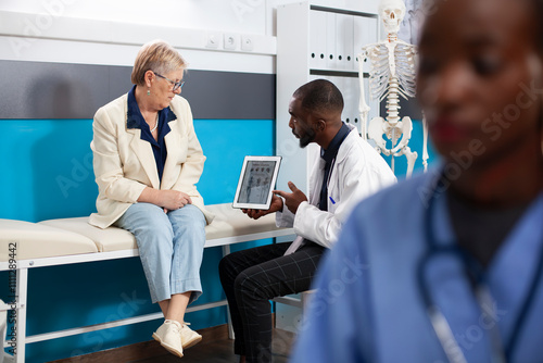 Black doctor explains healthcare treatment to caucasian old woman using diagrams on tablet. Selective focus on elderly patient and male physician reviewing medical information on digital device. photo