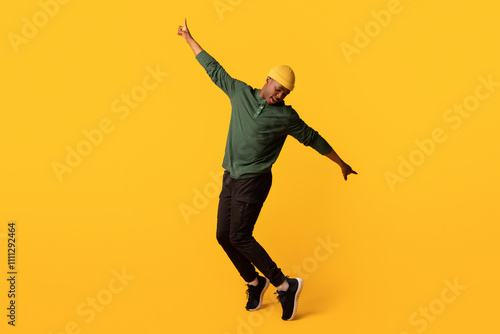 Cheerful young african american man in orange hat standing on tiptoes over yellow background, full length shot of happy millennial black hipster guy dancing and having fun in studio, copy space photo