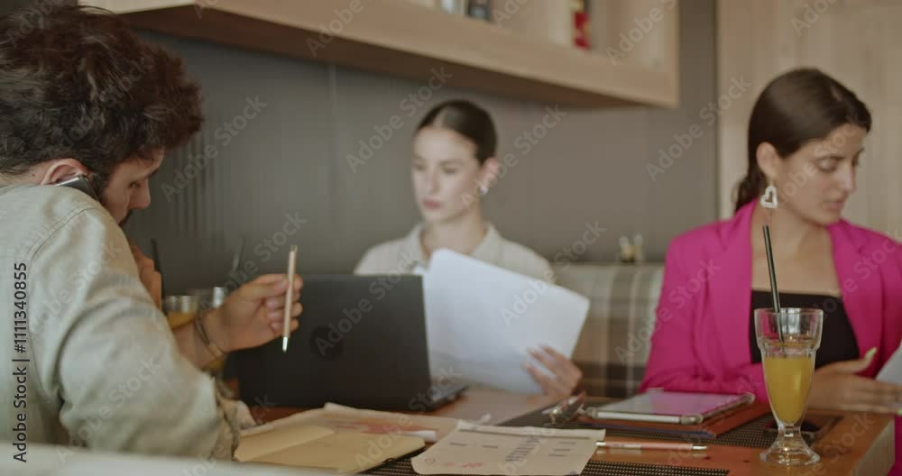 A business meeting unfolds outdoors on a summer day, where three colleagues collaborate around a table, working with papers, a laptop, and drinks as they discuss and strategize together. A business meeting unfolds outdoors on a summer day, where three colleagues collaborate around a table, working with papers, a laptop, and drinks as they discuss and strategize together.