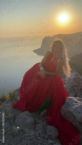 Beautiful blonde woman in a flowing red evening dress on the top of a cliff watches the sun set against the backdrop of the sea.