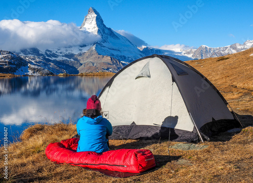 Person in a sleeping bag near a tent, admiring a stunning mountain peak - Matterhorn and lake on a sunny day. photo