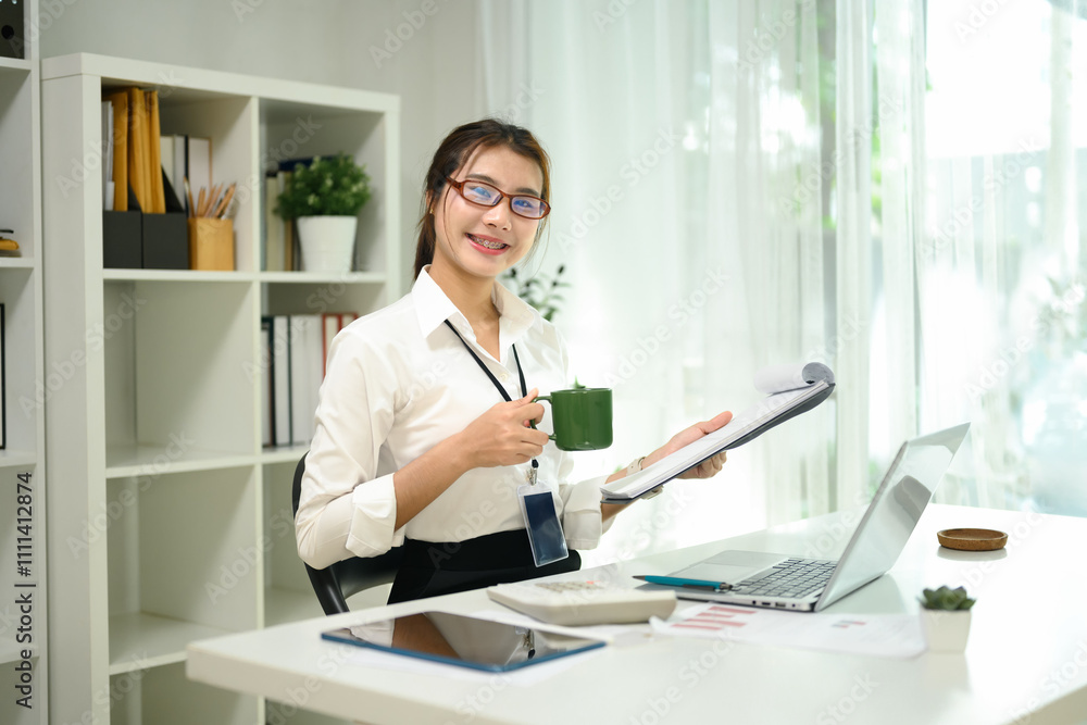 Cheerful asian female accountant holding coffee cup reviewing document at desk Cheerful asian female accountant holding coffee cup reviewing document at desk