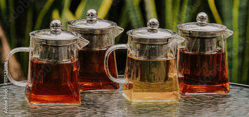 Fotografija A collection of clear teapots showcasing vibrant tea colors, elegantly arranged on a textured glass table against a natural green backdrop, creating a serene tea-drinking ambiance