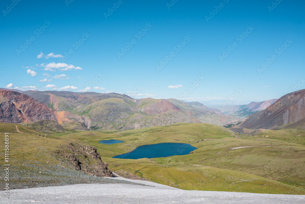 Top view from glacier to two beautiful blue lake among green hills in bright sun. Sunlit valley between multicolor mountains of many pastel colors in sunny day. Mountain range silhouette far away. Top view from glacier to two beautiful blue lake among green hills in bright sun. Sunlit valley between multicolor mountains of many pastel colors in sunny day. Mountain range silhouette far away.