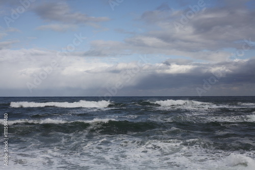 A sandy beach in Gangneung faces gentle waves, with a yellow beacon standing offshore. A faint rainbow arcs in the cloudy sky, adding charm to the serene seascape.