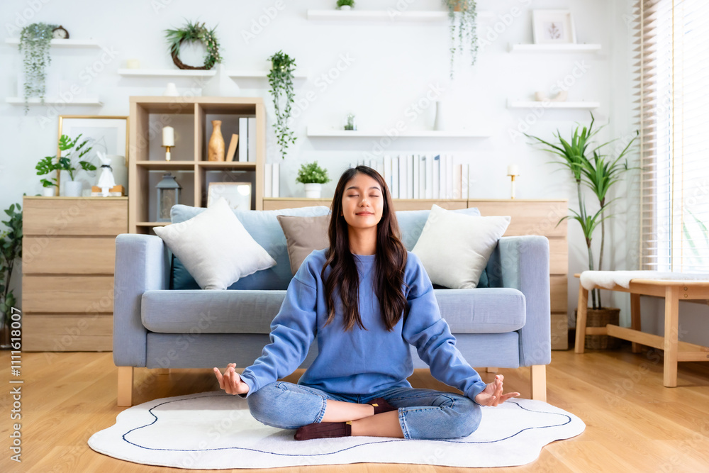 Happy young Asian woman practicing yoga and meditation at home sitting on floor in living room in lotus position and relaxing with closed eyes. Mindful meditation and wellbeing concept Happy young Asian woman practicing yoga and meditation at home sitting on floor in living room in lotus position and relaxing with closed eyes. Mindful meditation and wellbeing concept