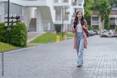 Portrait of a charming Asian female student in casual clothes. With backpack walking on campus her university Holding a book and smiling happily, enjoying the concept of people and education.