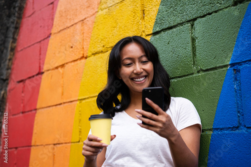 Happy woman using smart phone and leaning with coffee cup on multi colored wall photo