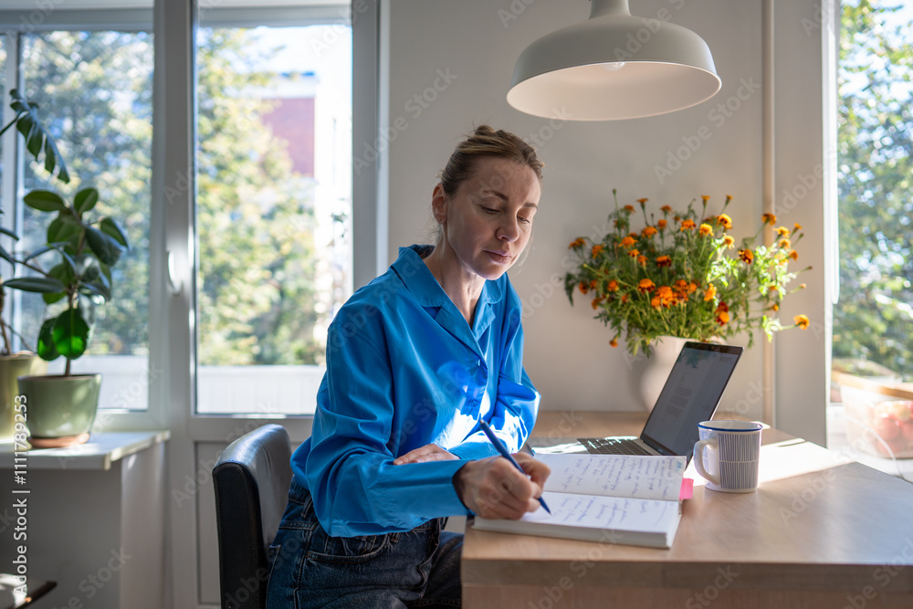 Φωτογραφία Concentrated middle aged woman works at home, planning day jotting down diary, comfortable sitting at table with laptop surround by houseplants