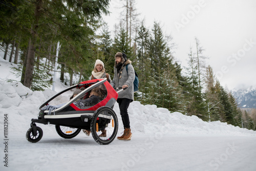 Young family is enjoying winter holiday in the mountains. Parents pushing kids in wagon stroller through snowy nature. photo