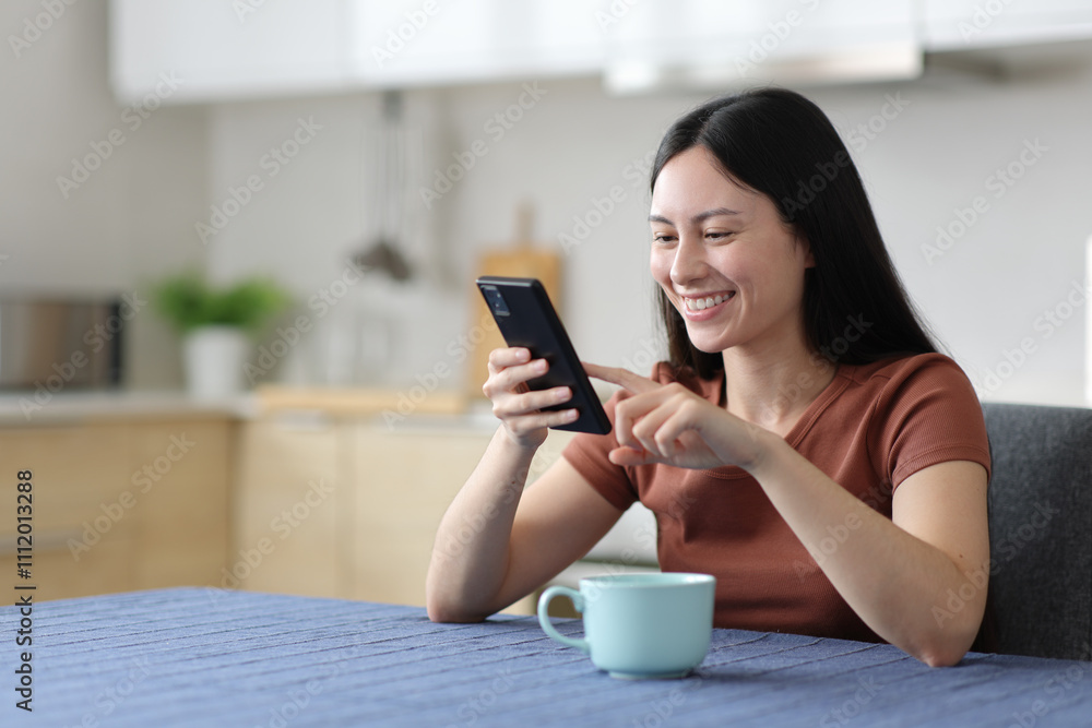 Happy asian woman checking cell phone in the kitchen Happy asian woman checking cell phone in the kitchen