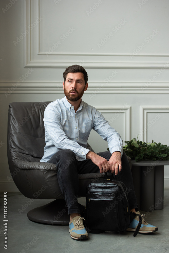 Fototapeta premium A man is comfortably sitting in a chair, resting with a leather backpack near him