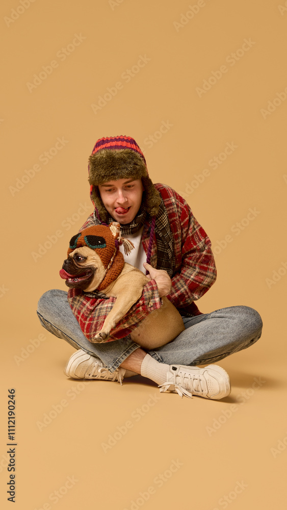 Playful cozy moment between young man in warm hat and checkered jacket, with Pug dog in knitted hat against studio background. Winter fun. Concept of animal, winter season, friendship, happiness Playful cozy moment between young man in warm hat and checkered jacket, with Pug dog in knitted hat against studio background. Winter fun. Concept of animal, winter season, friendship, happiness