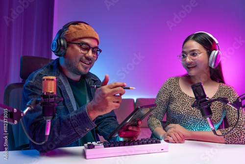 Two young asian streamer host making live podcast with condenser microphone and tablet at broadcast home studio. Content creator and social media influencer creating digital content.