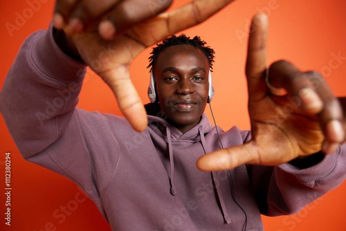 Smiling man wearing headphones and looking through finger frame against orange background photo