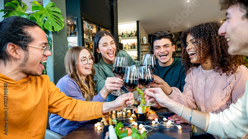 Group of multiracial friend toasting red wine sitting at bar restaurant table - Millennial people enjoying dinner party together - Life style concept with diverse guys and girls hanging out weekend