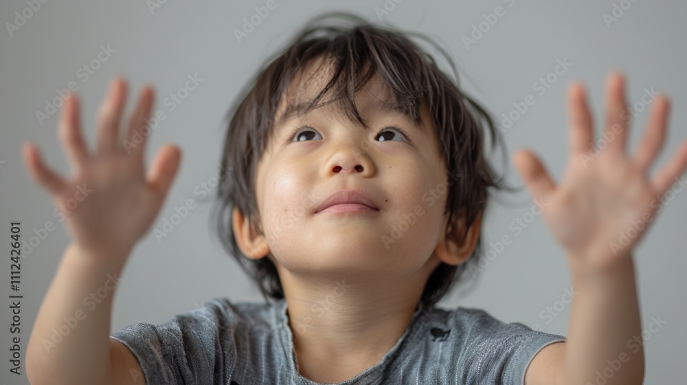 A young boy with dark hair and gray shirt looks up with his hands raised possibly reaching for something or expressing a gesture Of wonder or excitement. A young boy with dark hair and gray shirt looks up with his hands raised possibly reaching for something or expressing a gesture Of wonder or excitement.