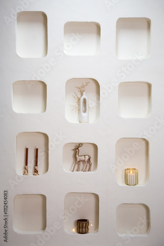 Minimalist wall display with built-in niches showcasing elegant holiday decor, including a white vase with delicate branches, metallic reindeer figurine, candles, and a glowing lantern.