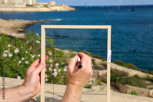 Strong male hands hold a weekly planner against the backdrop of the sea and rocky shores of Malta and write out a schedule for the next week.