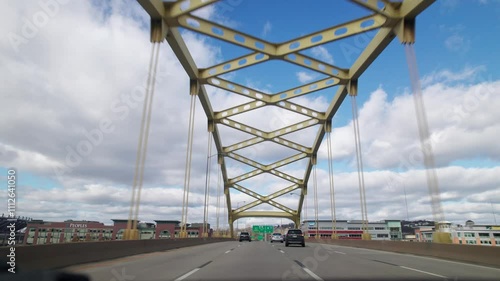 Car driver pov point of view moving toward Pittsburgh, PA city skyline and skyscraper buildings on multilane highway and interstate through vehicle traffic on golden bridge arches with blue sky
