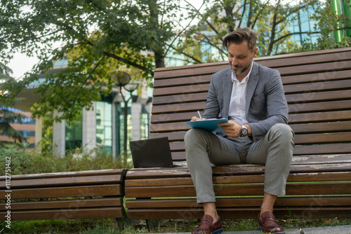 A business man writes on his notepad while sitting on a park bench, with modern glass office buildings in the background, blending work with the tranquility of the outdoors.