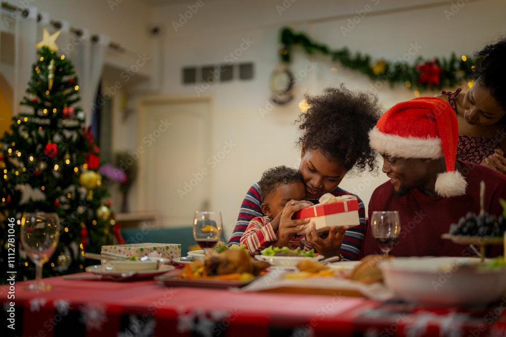 A joyful family gathers around the dinner table celebrating Christmas. The father, dressed in a Santa hat, gives a gift to the child, while everyone smiles with warmth and festive decorations. A joyful family gathers around the dinner table celebrating Christmas. The father, dressed in a Santa hat, gives a gift to the child, while everyone smiles with warmth and festive decorations.
