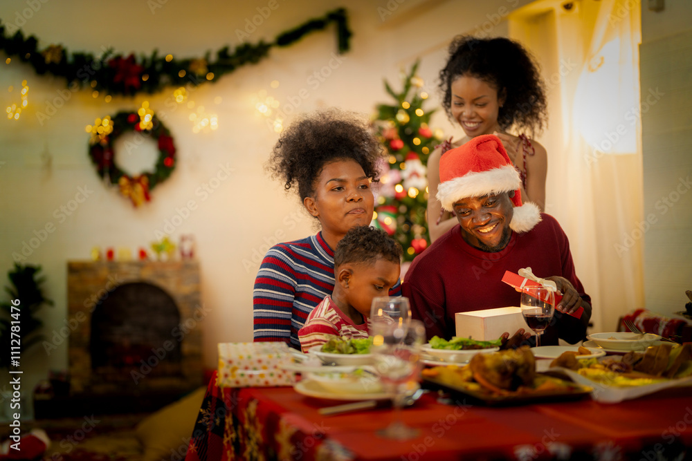 A joyful family gathers around the dinner table celebrating Christmas. The father, dressed in a Santa hat, gives a gift to the child, while everyone smiles with warmth and festive decorations. A joyful family gathers around the dinner table celebrating Christmas. The father, dressed in a Santa hat, gives a gift to the child, while everyone smiles with warmth and festive decorations.
