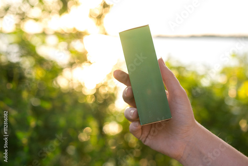 Hand holding a green rectangular box against a blurred natural background with sunlight. Perfect for eco-friendly product promotions and nature-themed advertisements.