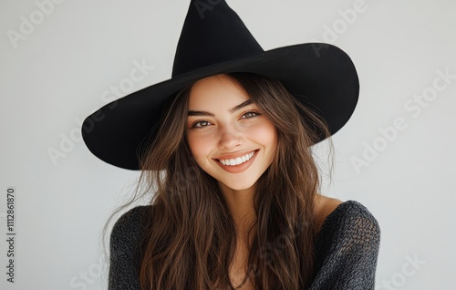 Smiling young woman with long hair and a large black hat posing against a lig...