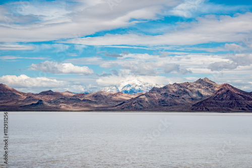 A snow-capped mountain with rugged foothills and a salt lake in the foreground under a partly cloudy sky. 