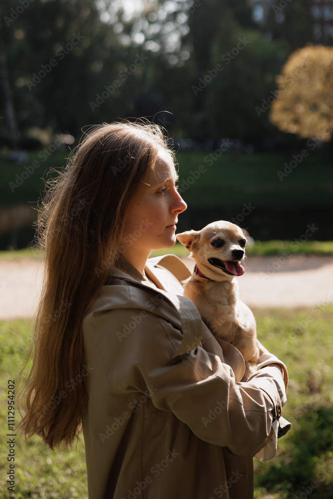 woman holds a small dog in her arms in park. dog is wearing a collar, and she is happy. lady is wearing beige raincoat and has long blonde hair. scene is peaceful and relaxing atmosphere woman holds a small dog in her arms in park. dog is wearing a collar, and she is happy. lady is wearing beige raincoat and has long blonde hair. scene is peaceful and relaxing atmosphere
