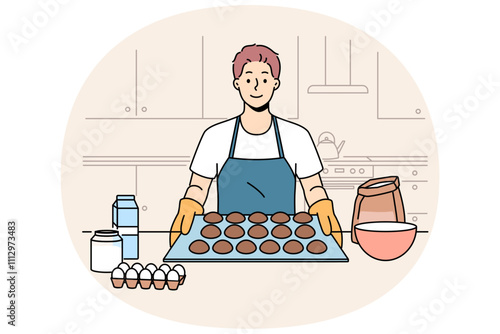Man baker prepares oatmeal cookies or gingerbread while standing near kitchen table with ingredients. Baker guy dressed in apron and gloves proudly demonstrates dessert ready for baking.
