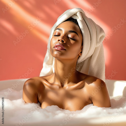  African-American woman in a bathtub with a white towel wrapped around her head, surrounded by bubble bath. Background of soft pink color. Spa treatments, cosmetology and body care. Relaxation practic photo