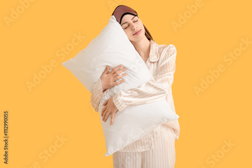 Young woman in pajamas and sleep mask with pillow on yellow background photo