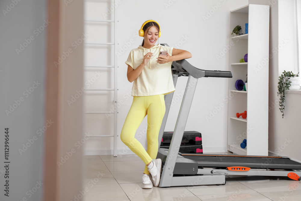 Fototapeta premium Sporty Asian woman in headphones with mobile phone near treadmill at gym